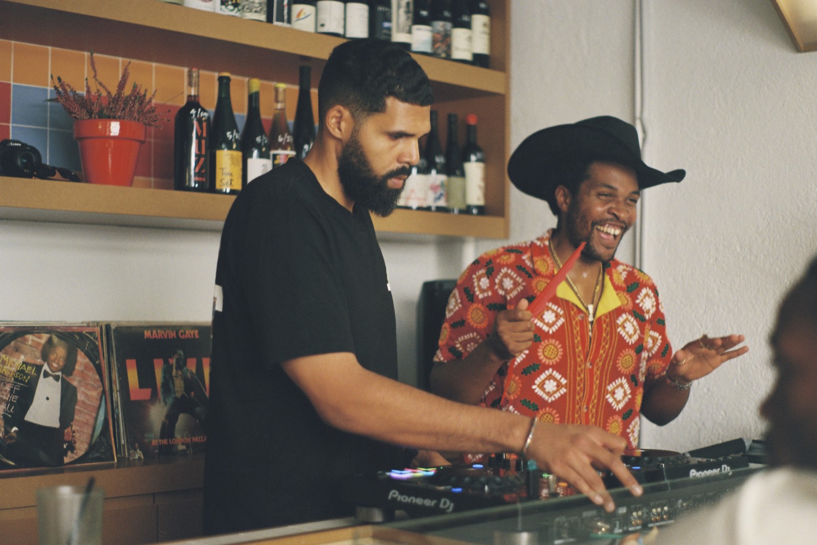 Two IFE SOUND selectors laughing behind the decks at LAZO Cafe and Wine Bar in Madrid, wine shelves and vinyl behind them, in full afternoon light